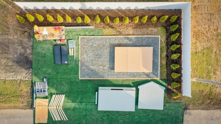 A bird’s-eye view of a landscaped backyard featuring a rectangular gravel area with a wooden platform, multiple small structures including sheds and storage units, and a neatly arranged row of evergreen trees along a white fence. The yard is divided into sections with artificial grass, a seating area, and a paved walkway leading to different areas.