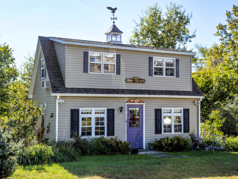 Two-story beige art studio shed with decorative shutters, a weather vane, and a garden in the foreground.