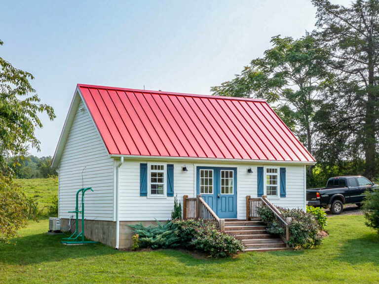 Small white home with a red metal roof, a covered porch, and flower beds in front, surrounded by a grassy field.