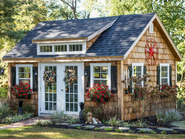 A picturesque cottage-style shed with natural cedar shake siding, a dark gray shingled roof, and white-trimmed French doors. Three transom windows above the doors allow natural light inside. Black shutters frame the front windows, adorned with vibrant red flower boxes. A decorative red metal star hangs on the side of the house. The landscape includes lush greenery, a stone walkway, and a small garden statue near the entrance.