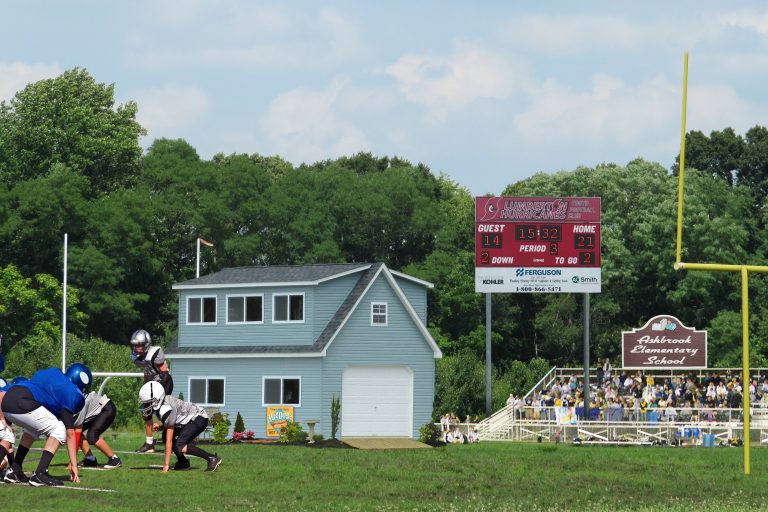A two-story blue storage shed with white trim and a gray shingled roof stands near a football field. The shed features multiple windows, a garage door on the lower level, and a small gable on the right side. A flag waves from a pole on the roof. In the foreground, two football players in black and white uniforms are engaged in play. Behind them, a scoreboard displays the current score, time, and period, showing the home team leading 21-14 in the third quarter. Spectators are seated in bleachers to the right, and lush green trees surround the scene under a partly cloudy sky.