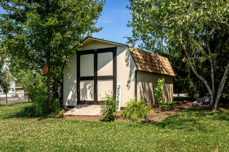 tan and black trimmed barn shed in between trees with ramp leading to double doors